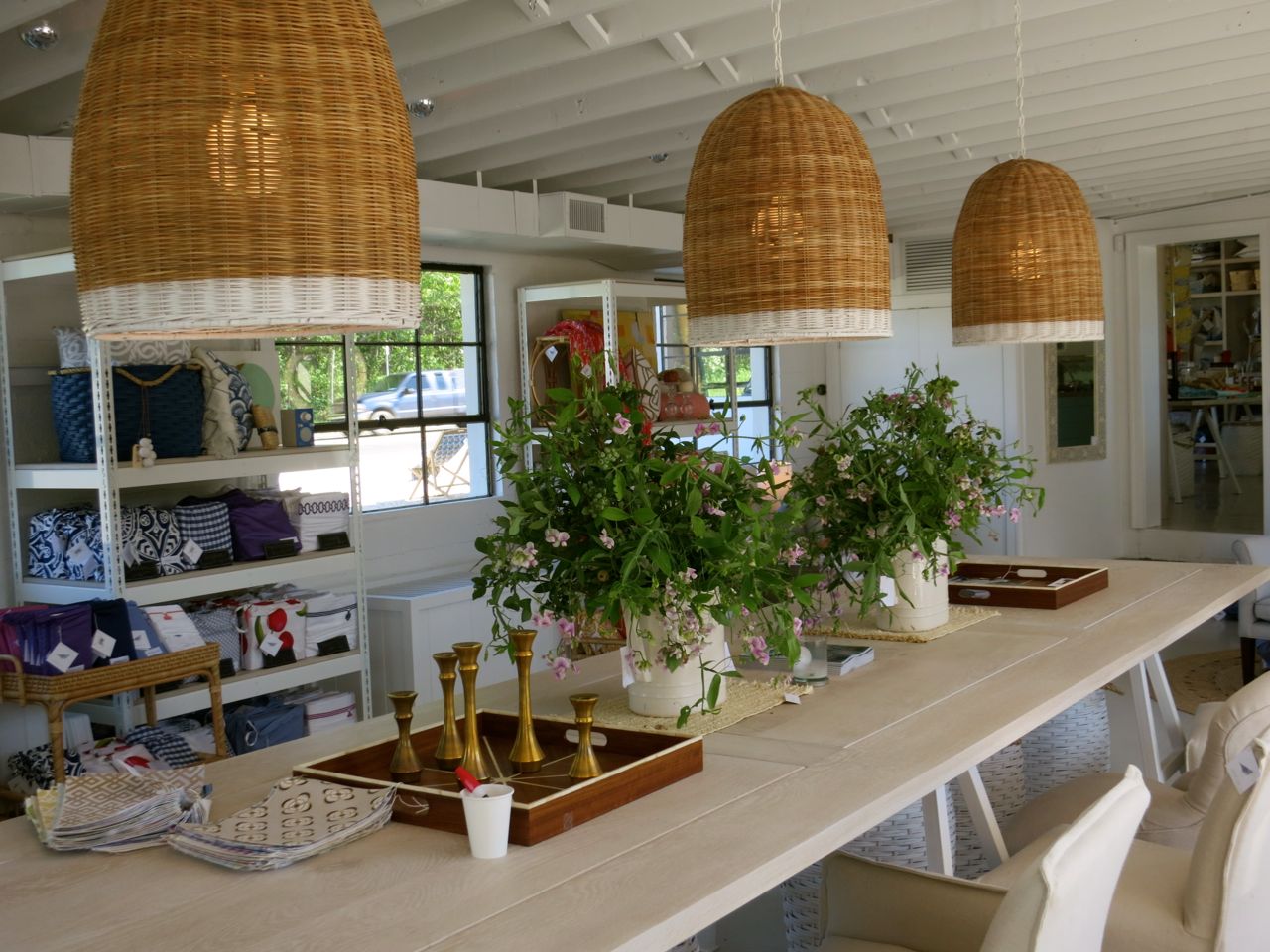 Shop interior with three large woven pendant lamps hanging over a long wooden table set with brass candlesticks and fresh greenery