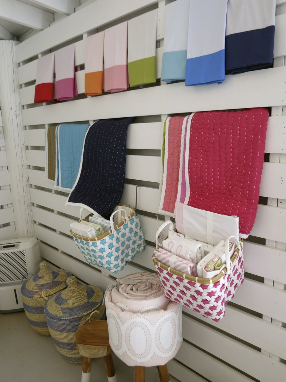 Colorful folded textiles and quilts displayed on a white slatted wall, with woven baskets and a small stool below