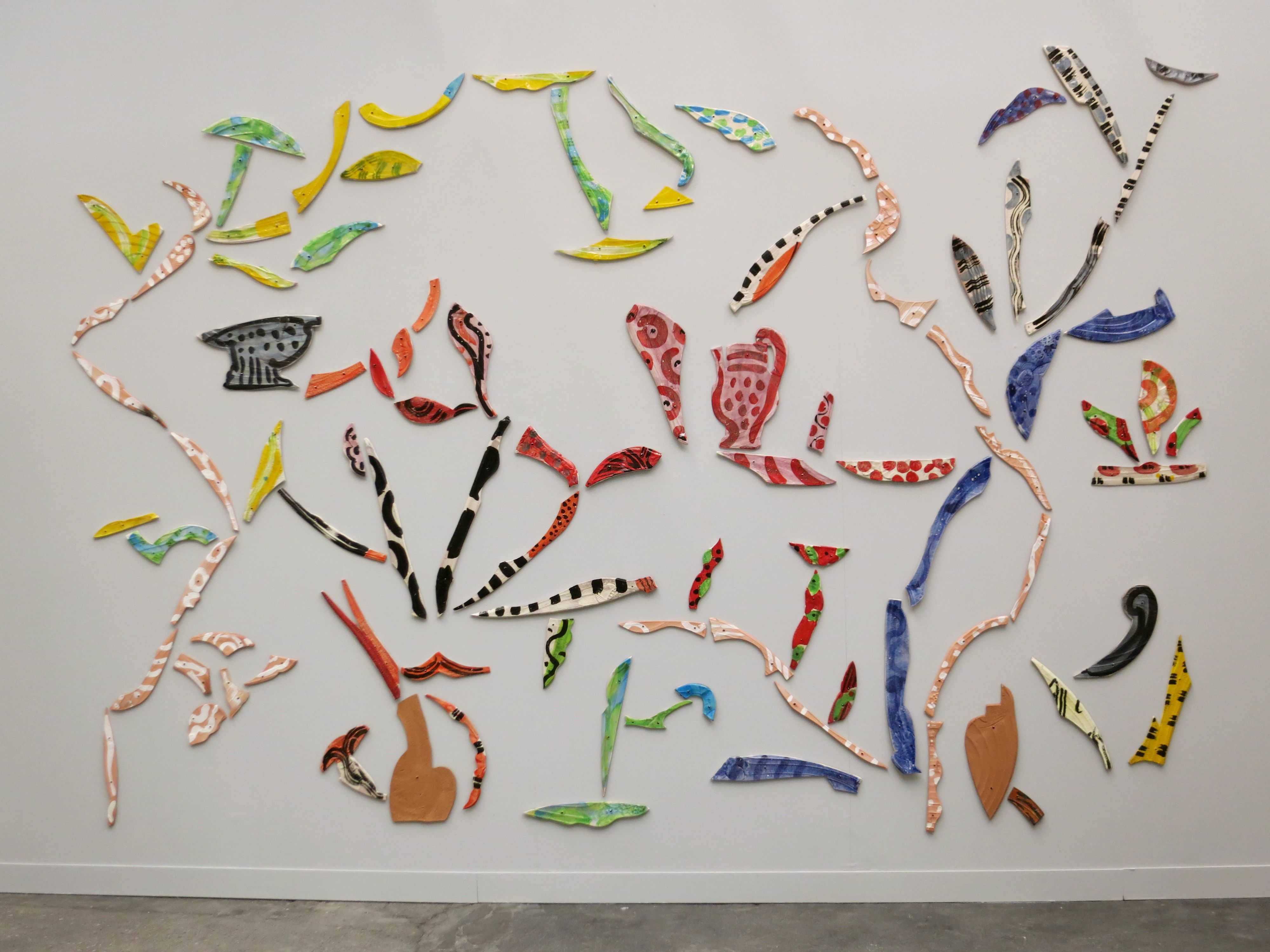 Wall installation of colorful painted ceramic sculptural fragments arranged in an abstract pattern against a light gray background