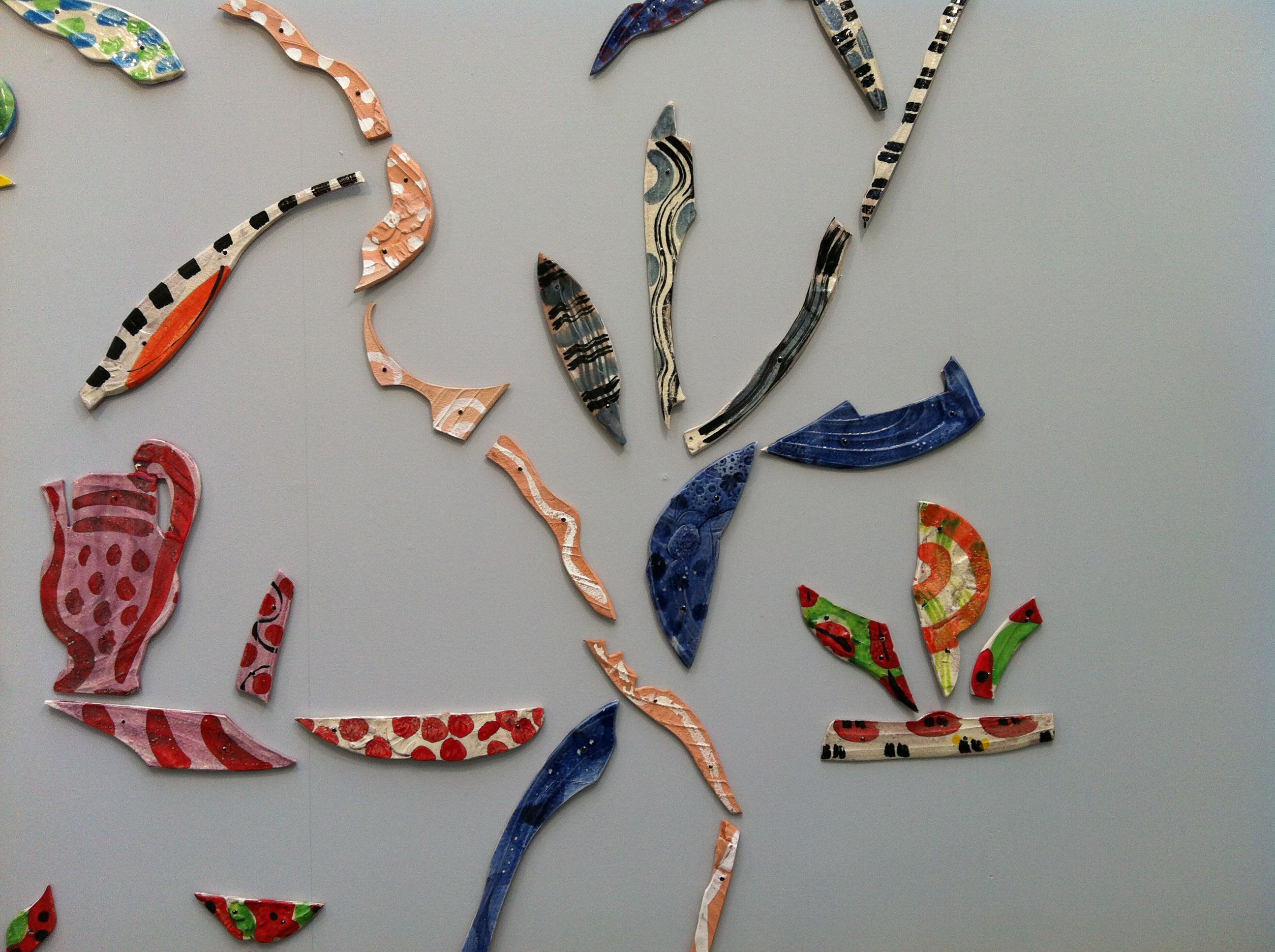 Detail of painted ceramic pieces including a jug shape and leaf-like forms in red, blue, and orange tones on a gray wall