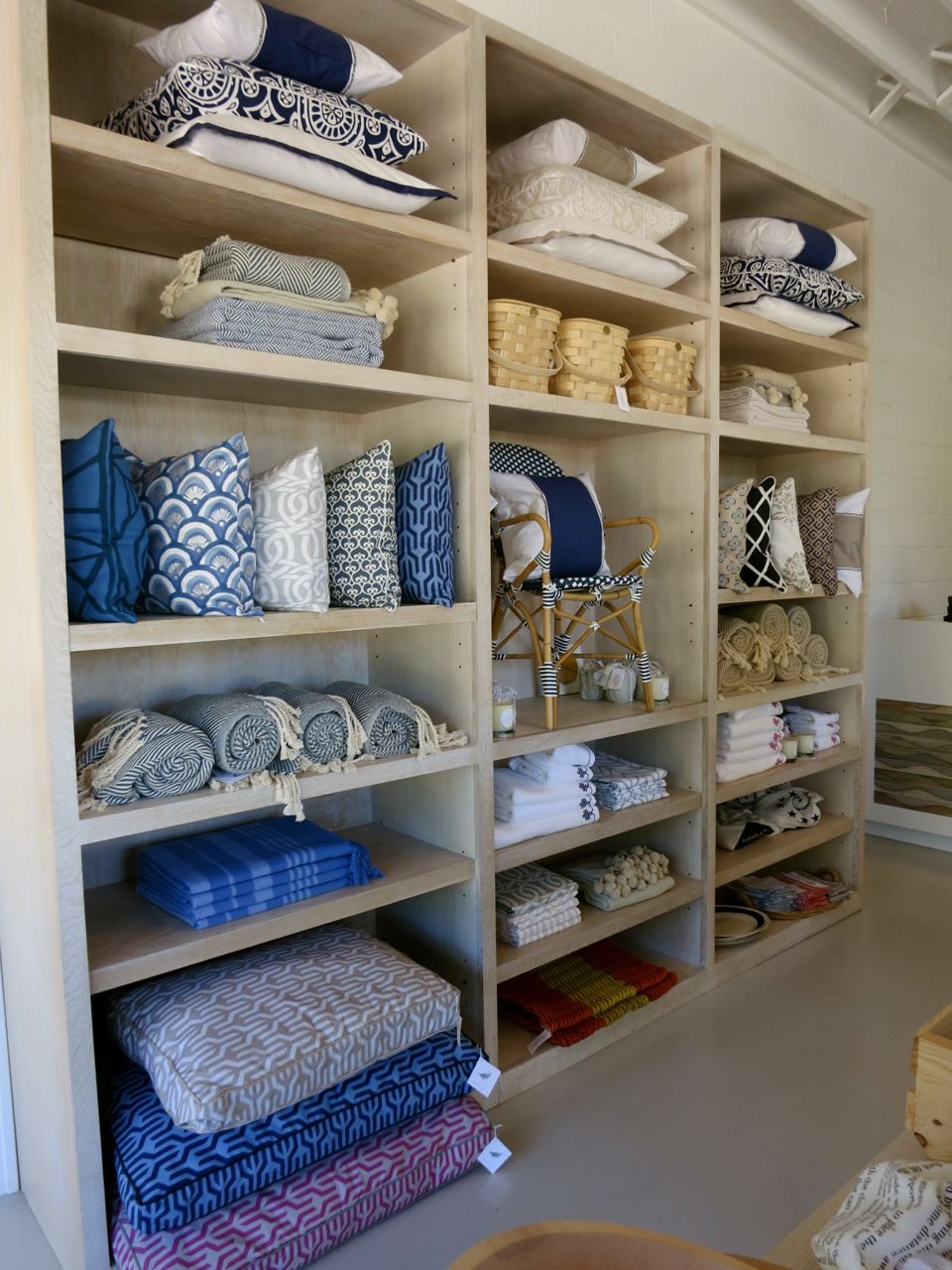 Wooden shelving filled with blue, white, and patterned pillows, folded towels, and baskets, neatly organized in a bright space